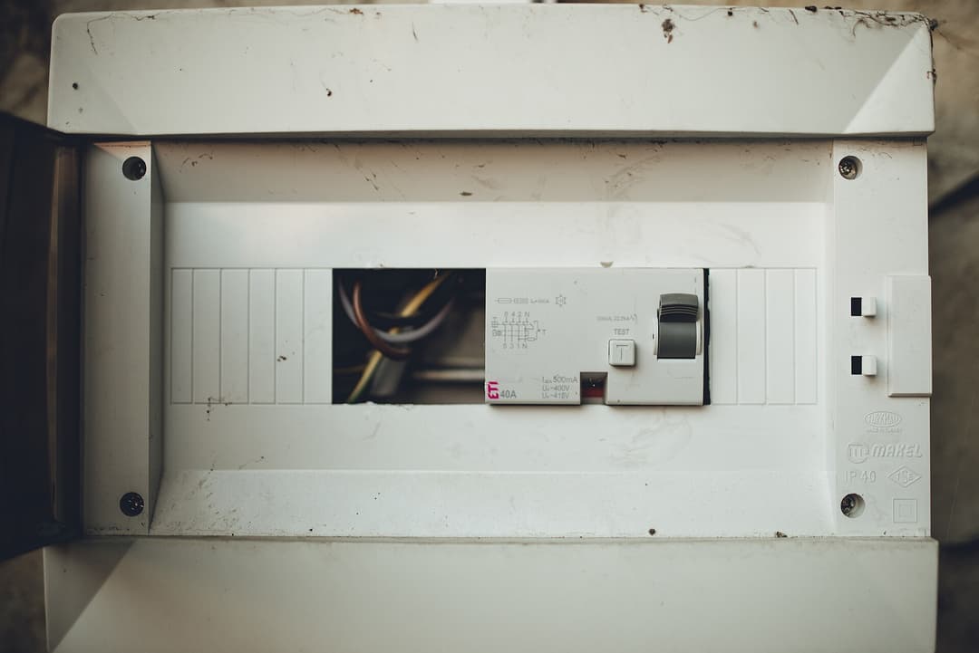 An electrical panel is shown with an open cover revealing wires and circuit breakers. The scene takes place in a home, with natural light coming in.